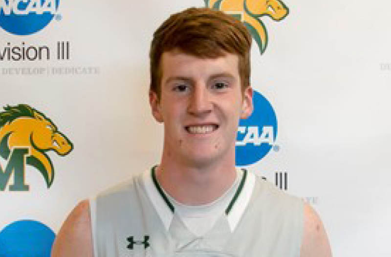 Smiling basketball player in a white jersey standing in front of a banner with a team logo and NCAA Division III logos.